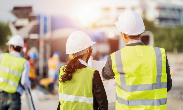 Twee bouwvakkers met witte helmen en gele vesten staan met hun rug naar de camera en bekijken plannen op een bouwplaats. Een wazige achtergrond toont meer arbeiders en apparatuur, met zonlicht dat de scène verlicht.