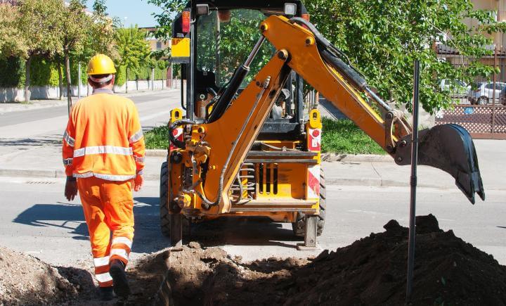 Een bouwvakker in oranje veiligheidskleding en een gele helm zorgt voor veilig werken bij een graaflaadmachine op een openbare weg. De machine graaft zich een weg in een berg aarde, met bomen en huizen als decor.