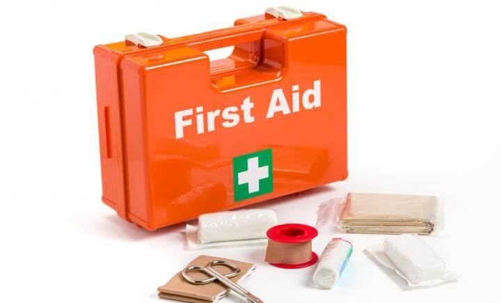 Een oranje EHBO-koffer met het label Eerste Hulp en een wit kruis wordt omringd door essentiële medische benodigdheden, waaronder scharen, verband, tape en gaasjes op een onberispelijk witte achtergrond.