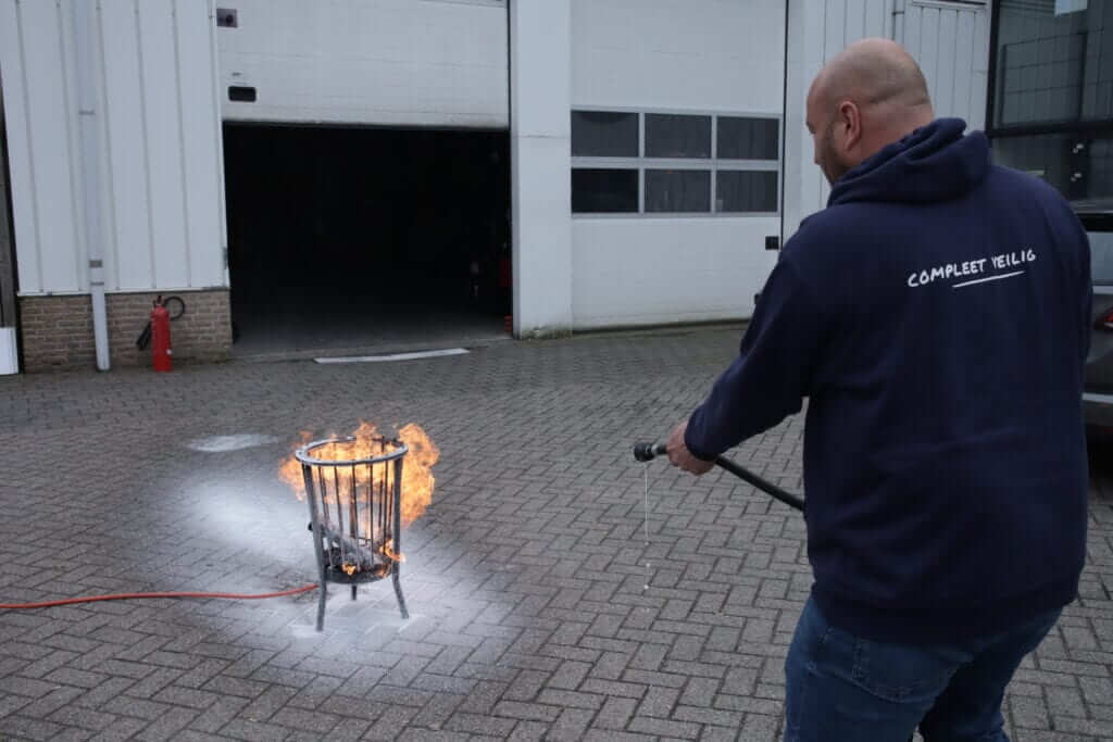 Een persoon in een donkere hoodie gebruikt een brandblusser om vlammen te doven in een metalen bak op het geplaveide oppervlak, waardoor brandveilige omstandigheden worden gegarandeerd. Een andere brandblusser is zichtbaar op de muur van het gebouw op de achtergrond.
