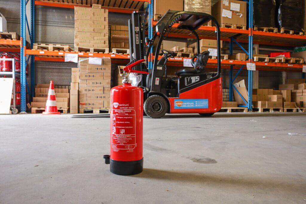 Een rode brandblusser, mogelijk een Welk-type brandblusser, staat op de vloer in een magazijn, met planken vol dozen en een rode vorkheftruck op de achtergrond. De scène lijkt georganiseerd en goed gevuld.
