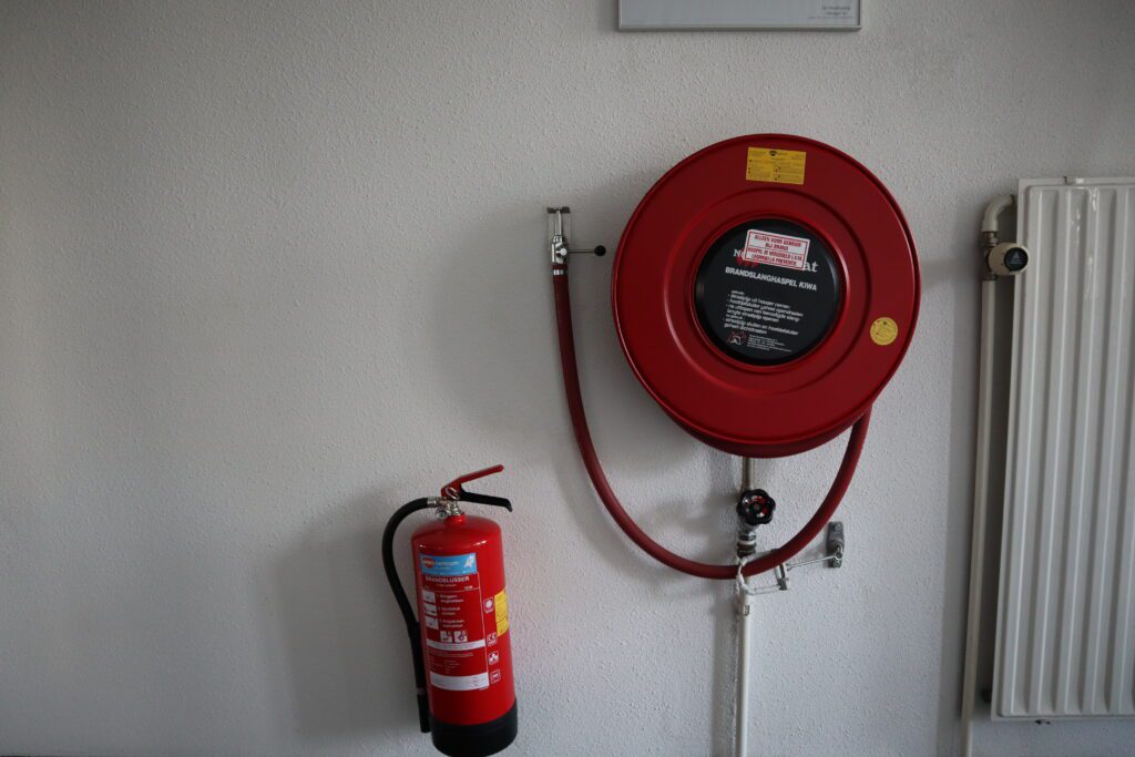 Een rode brandslanghaspel gemonteerd op een witte muur met daaronder een brandblusser benadrukt de veiligheidsvoorbereiding. Aan de rechterkant is een radiator zichtbaar, wat drie redenen onderstreept om een beheerder brandmeldinstallatie cursus te volgen: uitgebreide kennis, naleving en efficiënte respons.