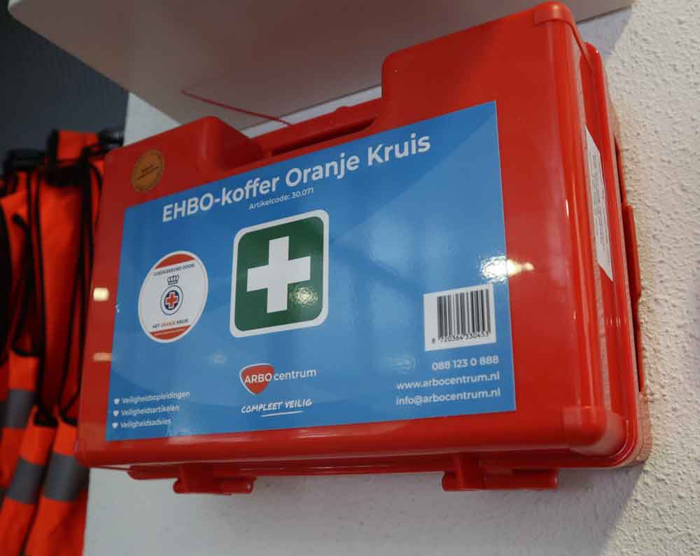 Een rode EHBO-doos met de opdruk EHBO-koffer Oranje Kruis is netjes aan de muur gemonteerd en onderstreept waarom een verbanddoos thuis aan te raden is. Op het etiket staan een groen kruis en contactgegevens. Er hangen felgekleurde veiligheidsvesten langszij, klaar voor elke noodsituatie.