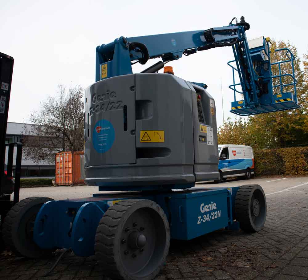 Een blauwe genie Z-34/22N hoogwerker staat geparkeerd op een verharde ondergrond buiten, met zijn giek uitgeschoven met een lege mand. Met bomen en een busje op de achtergrond, is het een herinnering aan de 5 redenen om een hoogwerkercertificaat te behalen voor veiligheid en bekwaamheid in het hanteren van dergelijke apparatuur.