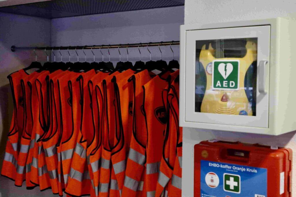 Een rij oranje veiligheidsvesten hangt netjes aan een rek in de kast. Naast de vesten staat een aan de muur bevestigde kast met daarin een AED-apparaat dat wordt onderhouden, en daaronder is een EHBO-doos zichtbaar.