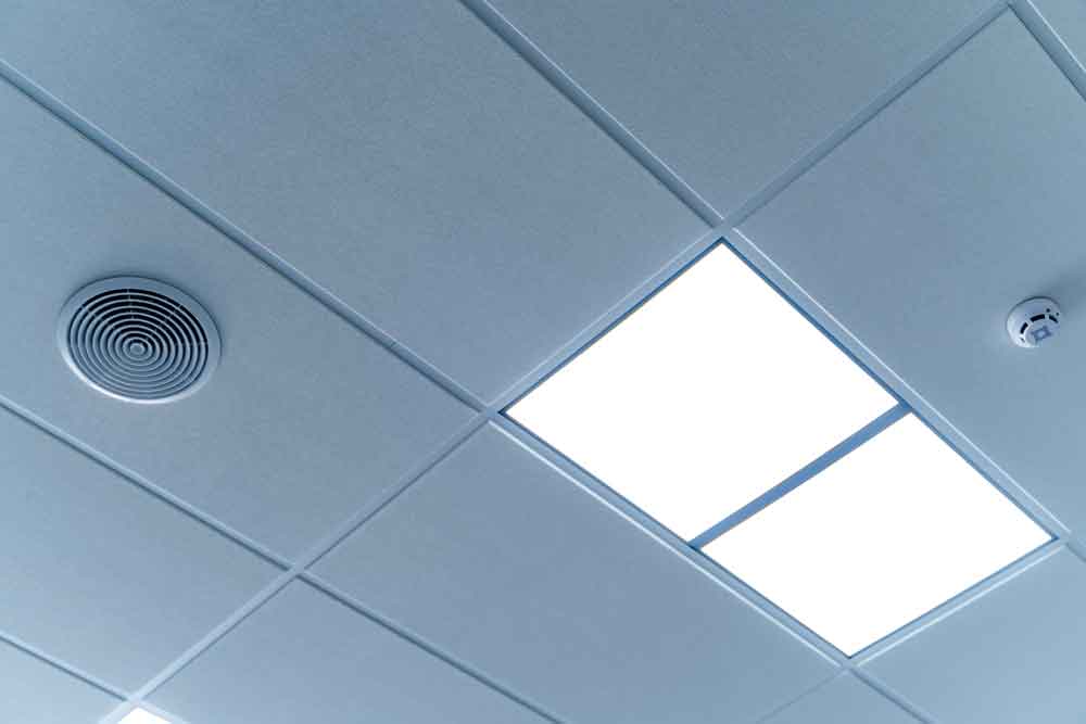 Een plafond met TL-verlichting, een ronde luchtopening en een kleine rookmelder. Met rechthoekige tegels in een rasterpatroon belichaamt het een van de 5 redenen om rookmelders te plaatsen door veiligheid en detectie-efficiëntie te garanderen.