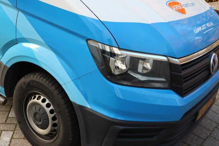 Close-up van de voorkant en zijkant van een blauwe bestelwagen, met een opvallende koplamp. De bestelwagen heeft branding met oranje accenten en tekst die gedeeltelijk zichtbaar is, wat misschien verwijst naar 4 stappen in het geval van autopech. Het voertuig staat geparkeerd op een geplaveide ondergrond.