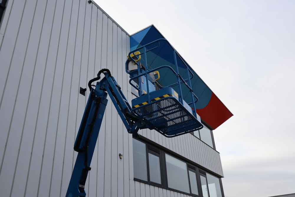 Een blauwe hoogwerkerlift staat naast een gebouw met witte verticale gevelbekleding en donkere ramen. Het platform van de lift bevindt zich bij een kleurrijke luifel boven de ramen, met blauwe en rode accenten. De lucht is bewolkt, wat illustreert waarom het behalen van een hoogwerkercertificaat essentieel is.