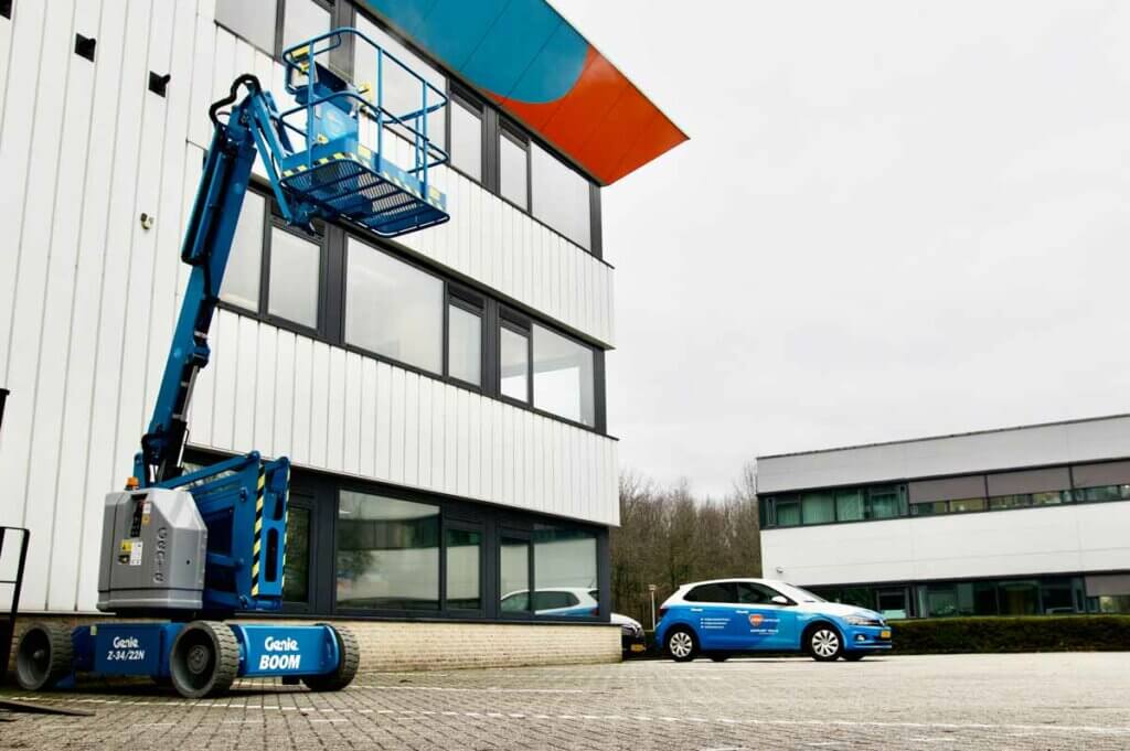 Een blauwe Genie-hoogwerker is opgetild bij de ramen van een wit industrieel gebouw, wat de voordelen van groepstraining in het bedienen van dergelijke machines illustreert. In de buurt staat een blauw-witte auto geparkeerd onder een bewolkte lucht, met een ander gebouw zichtbaar op de achtergrond.