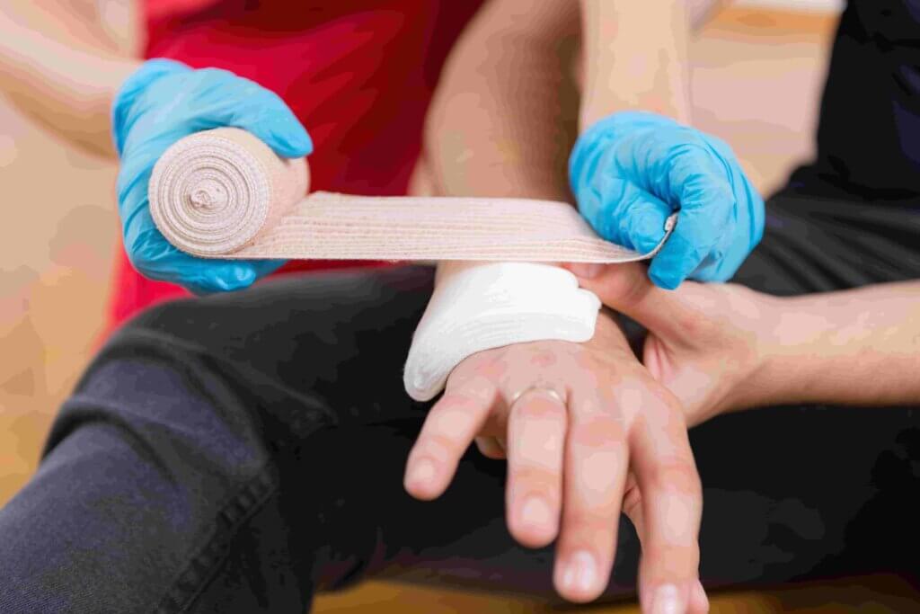 Een persoon die blauwe handschoenen draagt, wikkelt een verband om de pols en hand van een ander persoon. De gewonde hand rust op de knie van de zittende persoon. De achtergrond is zacht vervaagd.