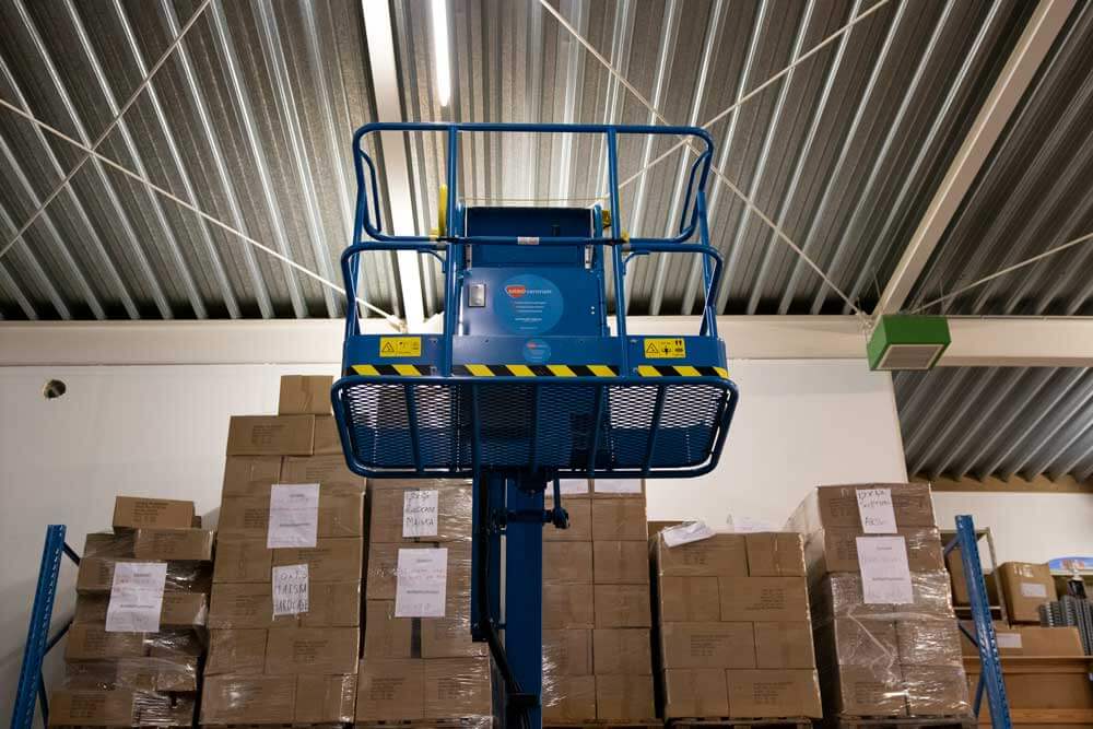 Een blauwe schaarlift wordt tussen hoge stapels kartonnen dozen gehesen in een magazijn met een metalen plafond. Aan de voorkant van sommige dozen zijn witte papieren bevestigd, misschien checklistnotities geïnspireerd door 3 tips om je voor te beginnen op de hoogwerker cursus. De lift is voorzien van gele en zwarte veiligheidsmarkeringen.