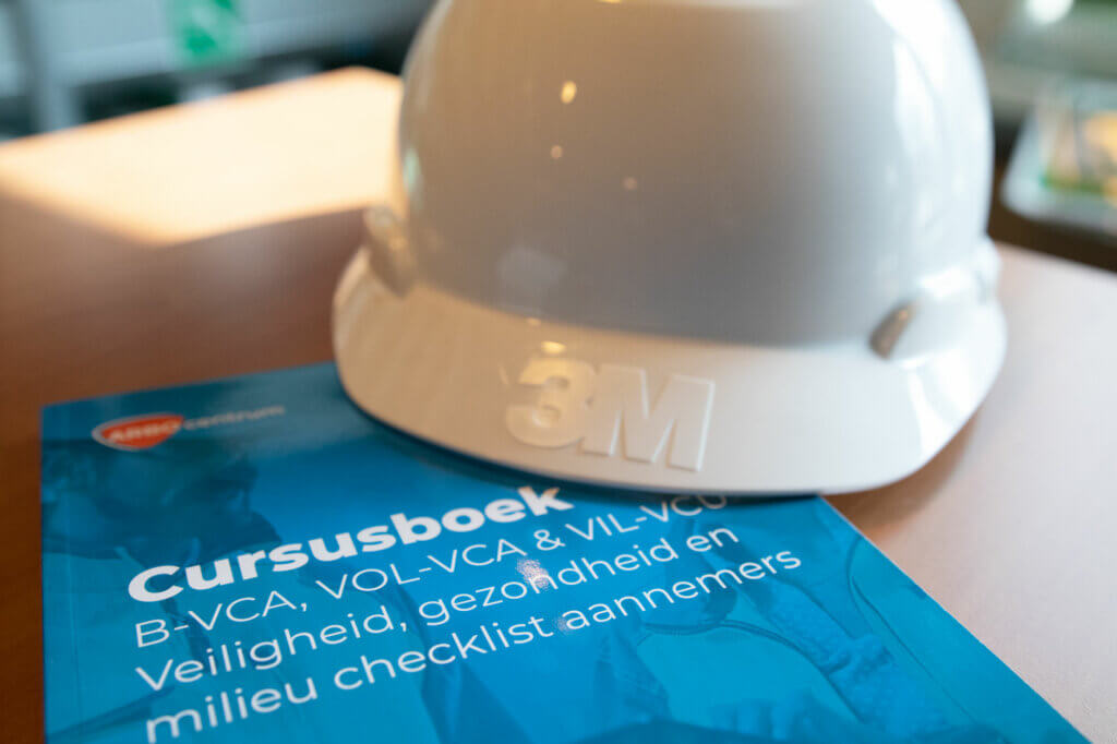 Op een tafel ligt een witte bouwvakker met het 3M-logo, naast een blauw cursusboek met de titel Cursusboek B-VCA, VOL-VCA & VIL-VCU veiligheid, gezondheid en milieu checklist aannemers, essentieel om te begrijpen hoe VCA hierbij wordt toegepast. sectoren.