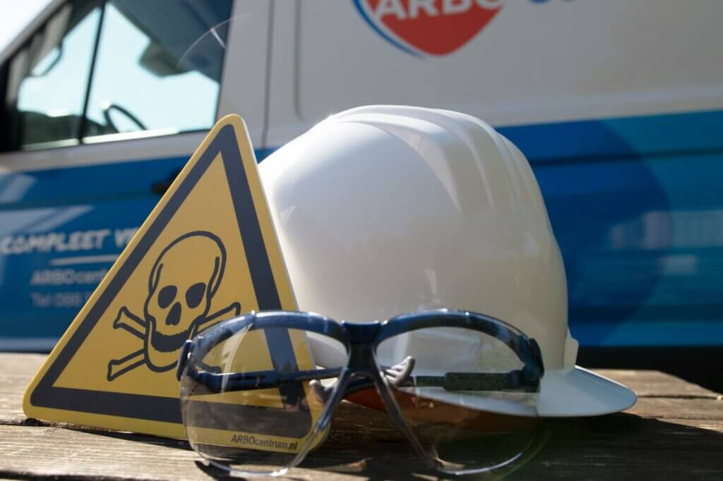 Een witte helm en veiligheidsbril rusten op een houten oppervlak, vergezeld door een geel driehoekig bord versierd met een zwarte doodshoofd en gekruiste beenderen. Op de achtergrond dient een blauw-wit voertuig als herinnering aan de 3 manieren om VCA te halen voor veiligheid op de werkplek.