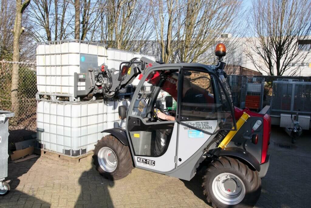 Een klein bouwvoertuig met een telescopische arm tilt een grote plastic container van een stapel buiten. De machine, die vaak wordt gebruikt in cursussen die 3 redenen om je verreiker certificaat te halen aanbieden, rijdt op een verhard oppervlak omgeven door bladerloze bomen en een hek van gaashekwerk.
