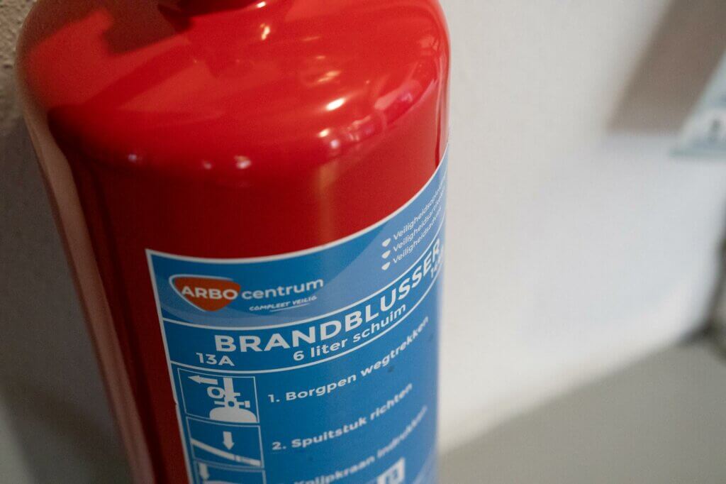 Close-up van een rode brandblusser die aan een muur is bevestigd. Het etiket in een vreemde taal geeft aan dat het een schuimblusser van 6 liter is, met instructies op het blauwe etiket. Dit dient als herinnering aan de redenen om je brandblusmiddelen jaarlijks te laten keuren voor veiligheid en naleving.