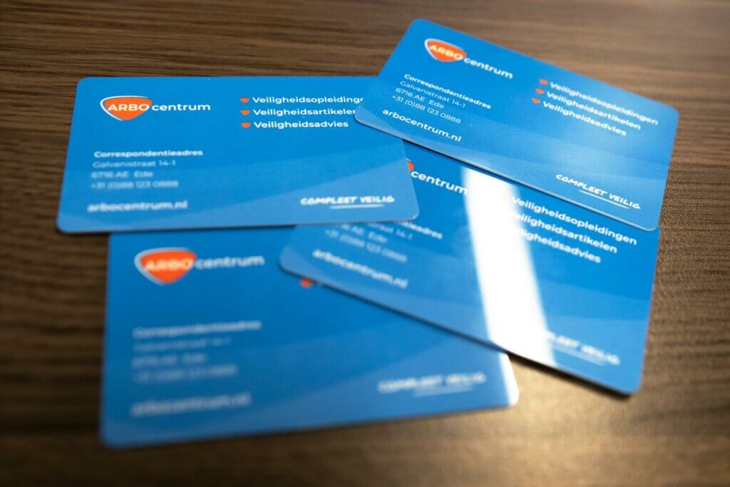 Vier blauwe visitekaartjes met oranje en witte tekst liggen verspreid op een houten oppervlak. De kaartjes, met de tekst Het nut van een VCA pas, hebben een logo in de linkerbovenhoek en contactgegevens in het Nederlands, inclusief de website-URL arbocentrum.nl.