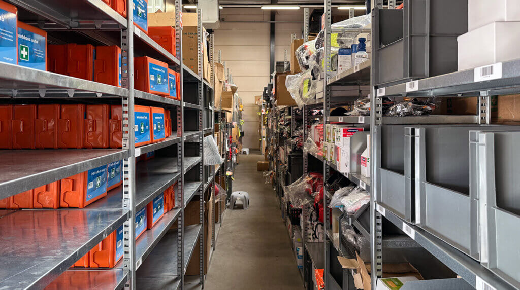 A cluttered warehouse aisle with metal shelves on both sides, filled with various boxes and items. Bright orange bins are visible on the left, and various packaged goods are stacked on the right. A concrete floor and overhead lights are visible.
