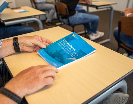 Close-up van een persoon die een blauwe gids met de titel Gas Book vasthoudt op een bureau in een klaslokaal. Andere studenten in casual kleding zitten aan lichte houten bureaus, sommigen houden soortgelijke boekjes vast. Deze scène is een voorbeeld van hoe de VCA-controle werkt in onderwijsomgevingen.